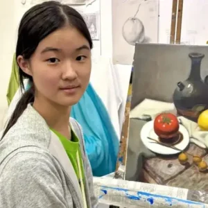 Student proudly showing her still life painting of a jug, tomato, lemon, and fruits on canvas at an art class easel