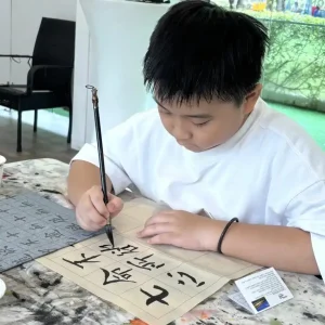 Young student practicing Chinese ink painting calligraphy, carefully brushing characters on grid paper with a traditional ink brush