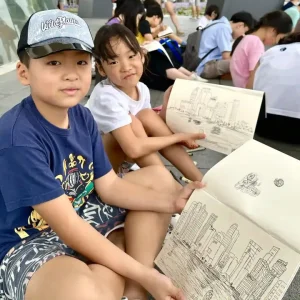 Two young students proudly showing their detailed urban sketching drawings of the city skyline, drawn during an outdoor sketching session